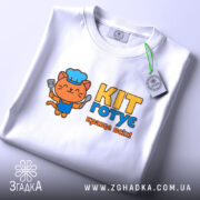 Футболка кіт готує, біла, з верхнього ракурсу, складена на поверхні, з яскравим принтом кота в кухарському ковпаку, що тримає лопатку, текстом 'KIT готує краще всіх!' у жовтому та блакитному кольорах, з етикеткою.