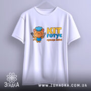 Футболка кіт готує, біла, з фронтального ракурсу, підвішена на вішалці, з яскравим принтом кота в кухарському ковпаку, що тримає лопатку, текстом 'KIT готує краще всіх!' у жовтому та блакитному кольорах.