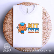 Футболка кіт готує, біла, з верхнього ракурсу, складена на плетеній основі, з яскравим принтом кота в кухарському ковпаку, що тримає лопатку, текстом 'KIT готує краще всіх!' у жовтому та блакитному кольорах.