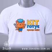 Футболка кіт готує, біла, з верхнього ракурсу, складена на поверхні, з яскравим принтом кота в кухарському ковпаку, що тримає лопатку, текстом 'KIT готує краще всіх!' у жовтому та блакитному кольорах, з етикеткою.