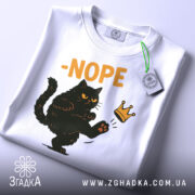Футболка кіт мем, біла, зображена з верхнього ракурсу, фон світлий, принт з чорним котом, що піднімає лапу, з написом '-NOPE' жовтого кольору, на футболці є етикетка з зеленим шнурком.