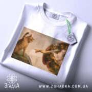 Футболка кіт Мікеланджело, біла, зображена на столі, фон світло-сірий, принт з котом і людиною в стилі Мікеланджело, на футболці є етикетка з зеленим шнурком, деталі малюнка чітко видимі.