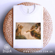 Футболка кіт Мікеланджело, біла, зображена на плетеній кошику, фон світло-сірий, принт з котом і людиною в стилі Мікеланджело, деталі малюнка чітко видимі.