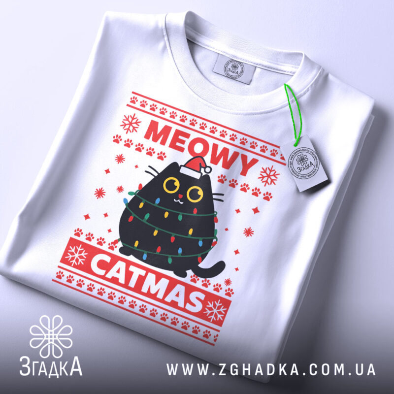 Футболка кіт на Різдво, біла, складена на поверхні, фон світлий, принт з чорним котом у різдвяних гірляндах і шапці Санти, текст 'MEOWY CATMAS' червоним кольором, з етикеткою на рукаві.