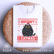 Футболка кіт на Різдво, біла, складена на плетеній основі, фон світлий, принт з чорним котом у різдвяних гірляндах і шапці Санти, текст 'MEOWY CATMAS' червоним кольором.