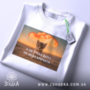Футболка кіт не треба кричати, біла футболка з принтом, котик на фоні вибуху, текст на українській мові, ракурс зверху, футболка складена, на ній зелена етикетка.