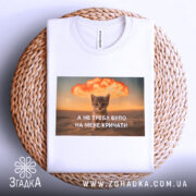 Футболка кіт не треба кричати, біла футболка з принтом, котик на фоні вибуху, текст на українській мові, ракурс зверху, футболка складена на плетеній основі.