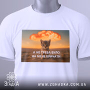 Футболка кіт не треба кричати, біла футболка з принтом, котик на фоні вибуху, текст на українській мові, ракурс зверху, футболка складена, на ній зелена етикетка.