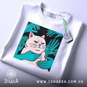 Футболка кіт незалежність, біла, складена на столі, фон світло-сірий, принт з котом та руками, що його обіймають, на етикетці зелена стрічка.