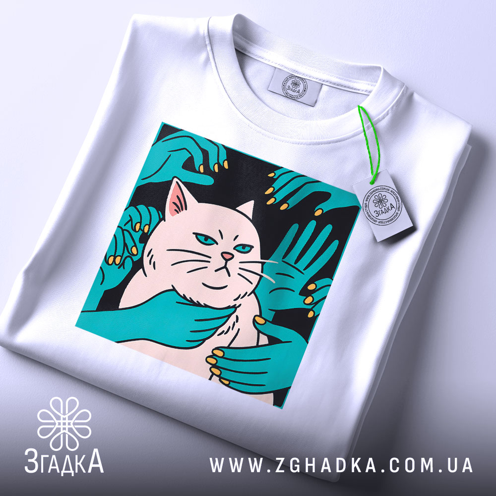 Футболка кіт незалежність, біла, складена на столі, фон світло-сірий, принт з котом та руками, що його обіймають, на етикетці зелена стрічка.