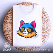 Футболка кіт патріот білого кольору, складена на плетеній кошику, ракурс зверху, фон світло-сірий, принт з котом у сонцезахисних окулярах з жовто-блакитним гербом України, видна етикетка на комірці.
