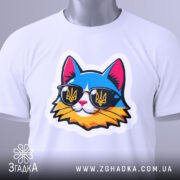 Футболка кіт патріот білого кольору, складена на поверхні, ракурс зверху, фон світло-сірий, принт з котом у сонцезахисних окулярах з жовто-блакитним гербом України, на комірці видна етикетка.