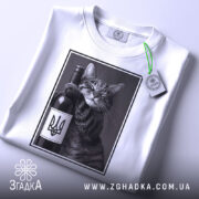 Футболка кіт патріот, біла, складена на столі, фон світло-сірий, принт з котом, що тримає пляшку вина з гербом України, на футболці є етикетка з логотипом.