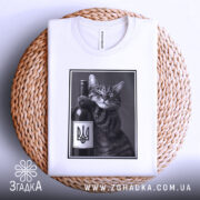 Футболка кіт патріот, біла, складена на плетеній кошику, фон світло-сірий, принт з котом, що тримає пляшку вина з гербом України, котяче обличчя з усмішкою.