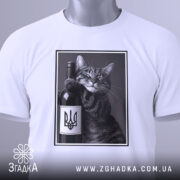 Футболка кіт патріот, біла, складена на столі, фон світло-сірий, принт з котом, що тримає пляшку вина з гербом України, на футболці є етикетка з логотипом.