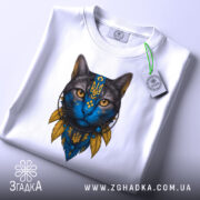 Футболка Кіт Патріот білого кольору, зображена на столі, ракурс зверху, фон світло-сірий, принт з котом з блакитним обличчям та жовтими деталями, на комірці видна етикетка.