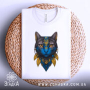 Футболка Кіт Патріот білого кольору, зображена на столі, ракурс зверху, фон світло-сірий, принт з котом з блакитним обличчям та жовтими деталями, футболка лежить на плетеній основі.