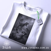 Футболка кіт проти динозавр, біла, складена на столі, фон світло-сірий, принт з котом, що лякається динозаврів, з етикеткою на рукаві.