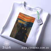 Футболка кіт стиль Мунка, біла, зображена на столі, фон світло-сірий, принт з чорним котом з великими жовтими очима, прикріплена етикетка з зеленим шнурком.