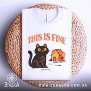 Футболка кіт this fine, складена біла футболка на плетеній основі, принт чорного кота на фоні палаючого будинку, ракурс зверху, фон світлий, текст 'THIS IS FINE' оранжевий.