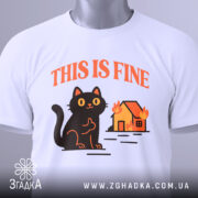 Футболка кіт this fine, складена біла футболка з принтом чорного кота, що сидить на фоні палаючого будинку, ракурс зверху, фон світлий, на футболці є етикетка з зеленим шнурком.