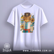 Футболка кіт у вишиванці, біла, підвішена на вішалці, фон світло-сірий, принт з котом у вишиванці, оточеним соняшниками та будинком.