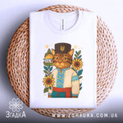 Футболка кіт у вишиванці, біла, складена на плетеній кошику, фон світло-сірий, принт з котом у вишиванці, оточеним соняшниками та будинком.