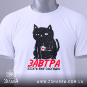 Футболка кіт з написом, біла, зображена з верхнього ракурсу, фон світлий, принт з чорним котом, який тримає чашку з серцем, текст червоним шрифтом 'ЗАВТРА БІСІТЬ ВЖЕ СЬОГОДНІ', на комірці видно етикетку.
