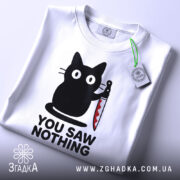 Футболка кіт з ножем, біла, складена на столі, фон світлий, принт з чорним котом, що тримає ніж, текст "YOU SAW NOTHING" чорним шрифтом під котом, на комірці є етикетка.