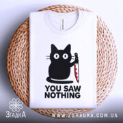 Футболка кіт з ножем, біла, складена на плетеній кошику, фон світлий, принт з чорним котом, що тримає ніж, текст "YOU SAW NOTHING" чорним шрифтом під котом.