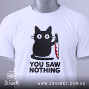 Футболка кіт з ножем, біла, складена на столі, фон світлий, принт з чорним котом, що тримає ніж, текст "YOU SAW NOTHING" чорним шрифтом під котом, на комірці є етикетка.