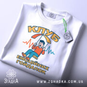 Футболка клуб емоційних гойдалок, біла, зображена з верхнього ракурсу, на фоні світло-сірого кольору, принт з хлопчиком на гойдалці, текст "КЛУБ ЕМОЦІЙНИХ ГОЙДАЛОК" в яскравих кольорах, з етикеткою на зеленій стрічці.