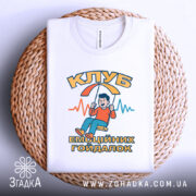 Футболка клуб емоційних гойдалок, біла, зображена зверху на плетеній кошику, на фоні світло-сірого кольору, принт з хлопчиком на гойдалці, текст "КЛУБ ЕМОЦІЙНИХ ГОЙДАЛОК" в яскравих кольорах.