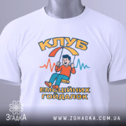 Футболка клуб емоційних гойдалок, біла, зображена з верхнього ракурсу, на фоні світло-сірого кольору, принт з хлопчиком на гойдалці, текст "КЛУБ ЕМОЦІЙНИХ ГОЙДАЛОК" в яскравих кольорах, з етикеткою на зеленій стрічці.