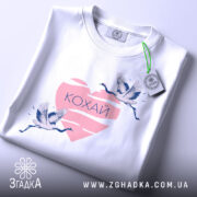 Футболка кохай птахи, біла, зображена зверху, на фоні світло-сірого кольору, принт з двома журавлями, що летять, на рожевому серці з написом 'Кохай', з етикеткою зеленого кольору.