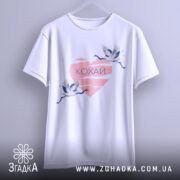 Футболка кохай птахи, біла, зображена на вішалці, на фоні світло-сірого кольору, принт з двома журавлями, що летять, на рожевому серці з написом 'Кохай'.