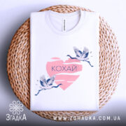 Футболка кохай птахи, біла, зображена зверху на плетеній основі, на фоні світло-сірого кольору, принт з двома журавлями, що летять, на рожевому серці з написом 'Кохай'.