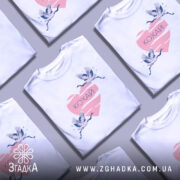 Футболка кохай птахи, біла, зображена з правого боку, на фоні світло-сірого кольору, принт з двома журавлями, що летять, на рожевому серці з написом 'Кохай'.