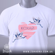 Футболка кохай птахи, біла, зображена зверху, на фоні світло-сірого кольору, принт з двома журавлями, що летять, на рожевому серці з написом 'Кохай', з етикеткою зеленого кольору.