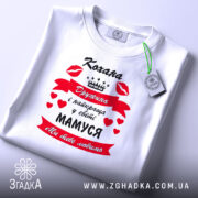 Футболка кохана дружина, біла, зображена зверху, на столі, фон білий, принт червоний з чорним текстом і сердечками, напис "Кохана Дружина і найкраща у світі МАМУСЯ Ми тебе любимо", з зеленим ярликом.