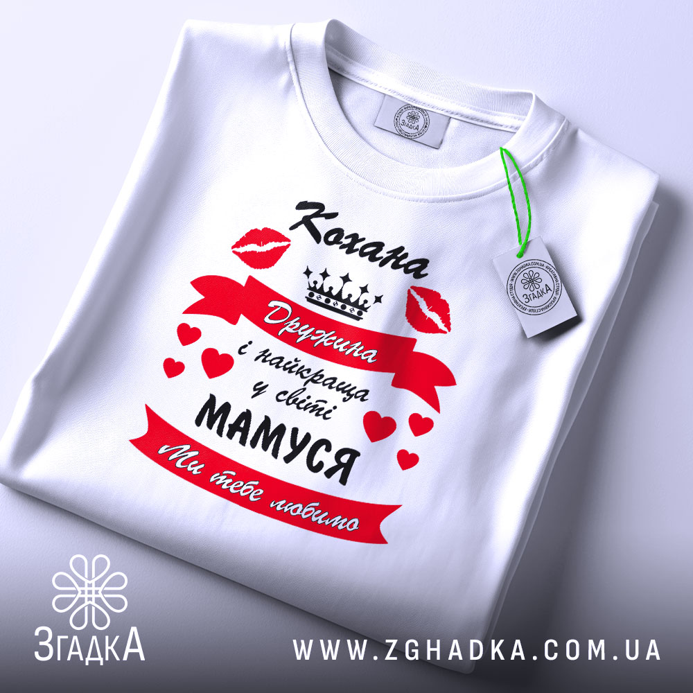 Футболка кохана дружина, біла, зображена зверху, на столі, фон білий, принт червоний з чорним текстом і сердечками, напис "Кохана Дружина і найкраща у світі МАМУСЯ Ми тебе любимо", з зеленим ярликом.