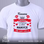 Футболка кохана дружина, біла, зображена зверху, на столі, фон білий, принт червоний з чорним текстом і сердечками, напис "Кохана Дружина і найкраща у світі МАМУСЯ Ми тебе любимо", з зеленим ярликом.