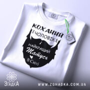 Футболка коханий чоловік, біла, складена на поверхні, фон світло-сірий, принт чорного кольору з написом 'Коханий чоловік і найкращий татусь в світі', з зеленим ярликом.