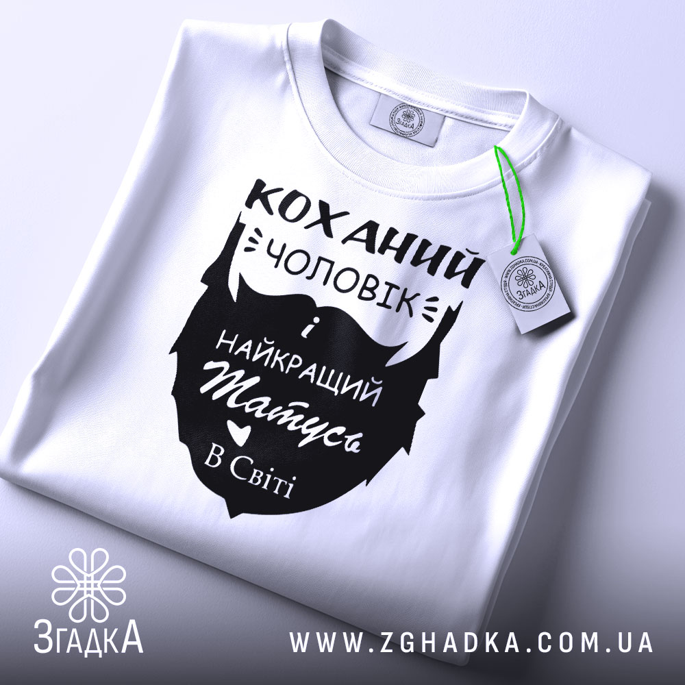 Футболка коханий чоловік, біла, складена на поверхні, фон світло-сірий, принт чорного кольору з написом 'Коханий чоловік і найкращий татусь в світі', з зеленим ярликом.