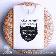 Футболка коханий чоловік, біла, складена на плетеній кошику, фон світло-сірий, принт чорного кольору з написом 'Коханий чоловік і найкращий татусь в світі'.