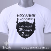 Футболка коханий чоловік, біла, складена на поверхні, фон світло-сірий, принт чорного кольору з написом 'Коханий чоловік і найкращий татусь в світі', з зеленим ярликом.