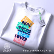 Футболка кольорові касети, біла, зображена зверху, на столі, фон світлий, принт з кольоровими касетами та написом 'REAL MUSIC LOVER' у темно-синьому кольорі, на футболці є етикетка.