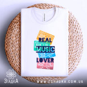 Футболка кольорові касети, біла, зображена зверху на плетеному кошику, фон світлий, принт з кольоровими касетами та написом 'REAL MUSIC LOVER' у темно-синьому кольорі.