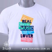 Футболка кольорові касети, біла, зображена зверху, на столі, фон світлий, принт з кольоровими касетами та написом 'REAL MUSIC LOVER' у темно-синьому кольорі, на футболці є етикетка.