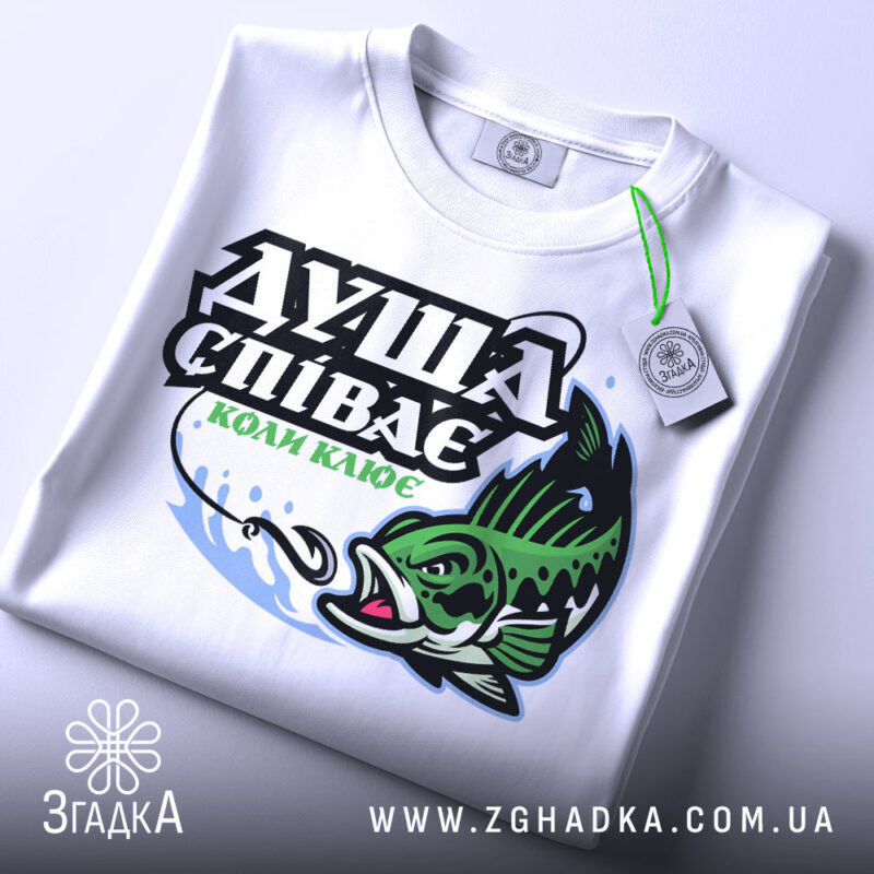 Футболка коли клює, біла, складена на поверхні, фон світло-сірий, принт з рибою та текстом "ДУША СПІВАЄ КОЛИ КЛЮЄ" в зелених та чорних кольорах, з етикеткою на шнурку.